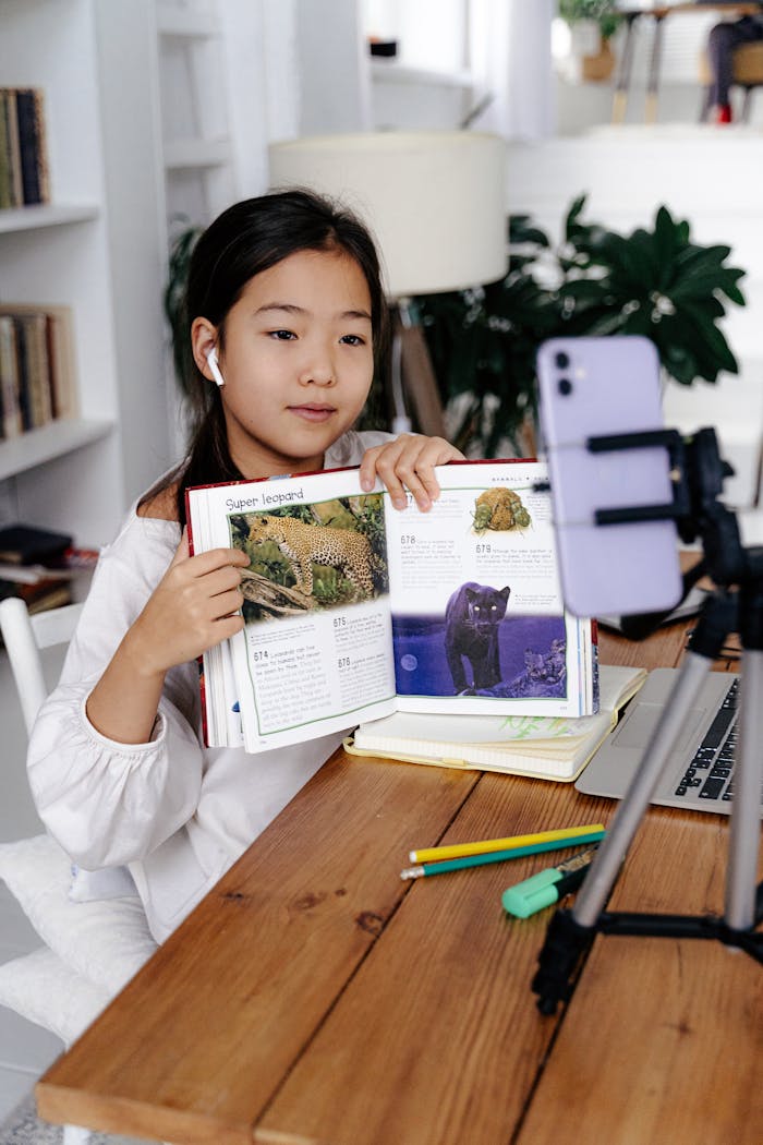 Young girl displays book during online class using smartphone and tripod.
