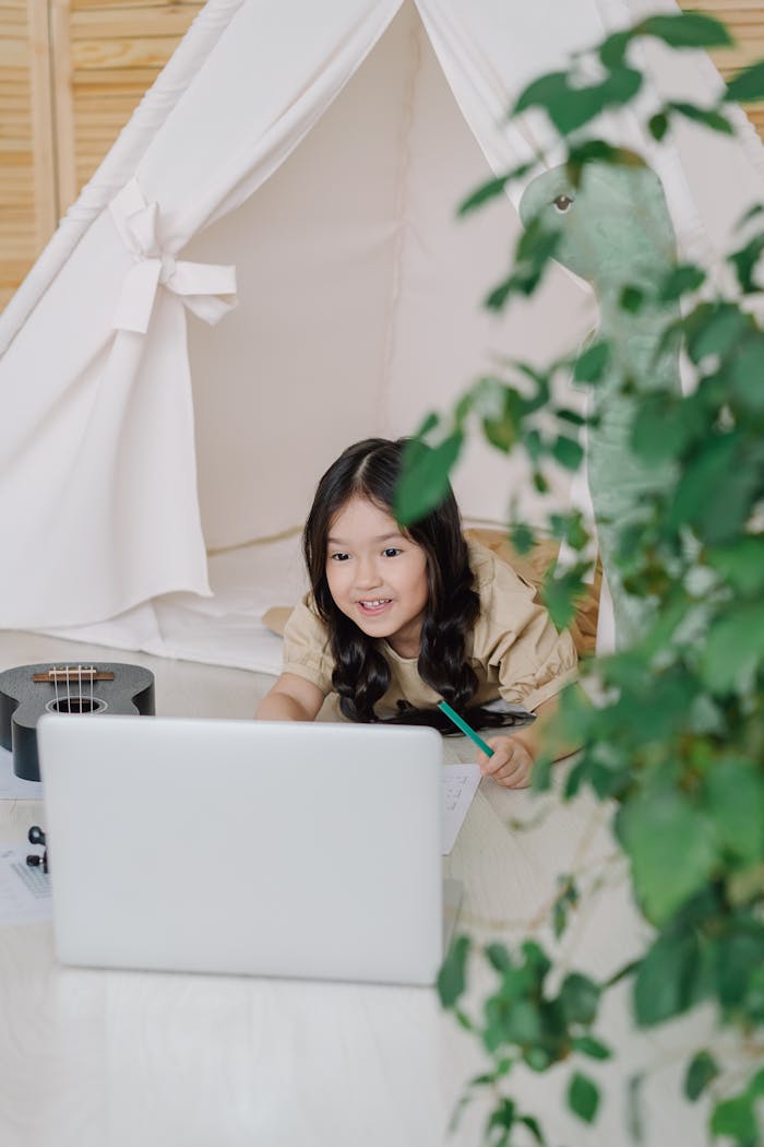 services-04 A young girl smiles while interacting with a laptop inside a cozy indoor tent, surrounded by a relaxing home setting.